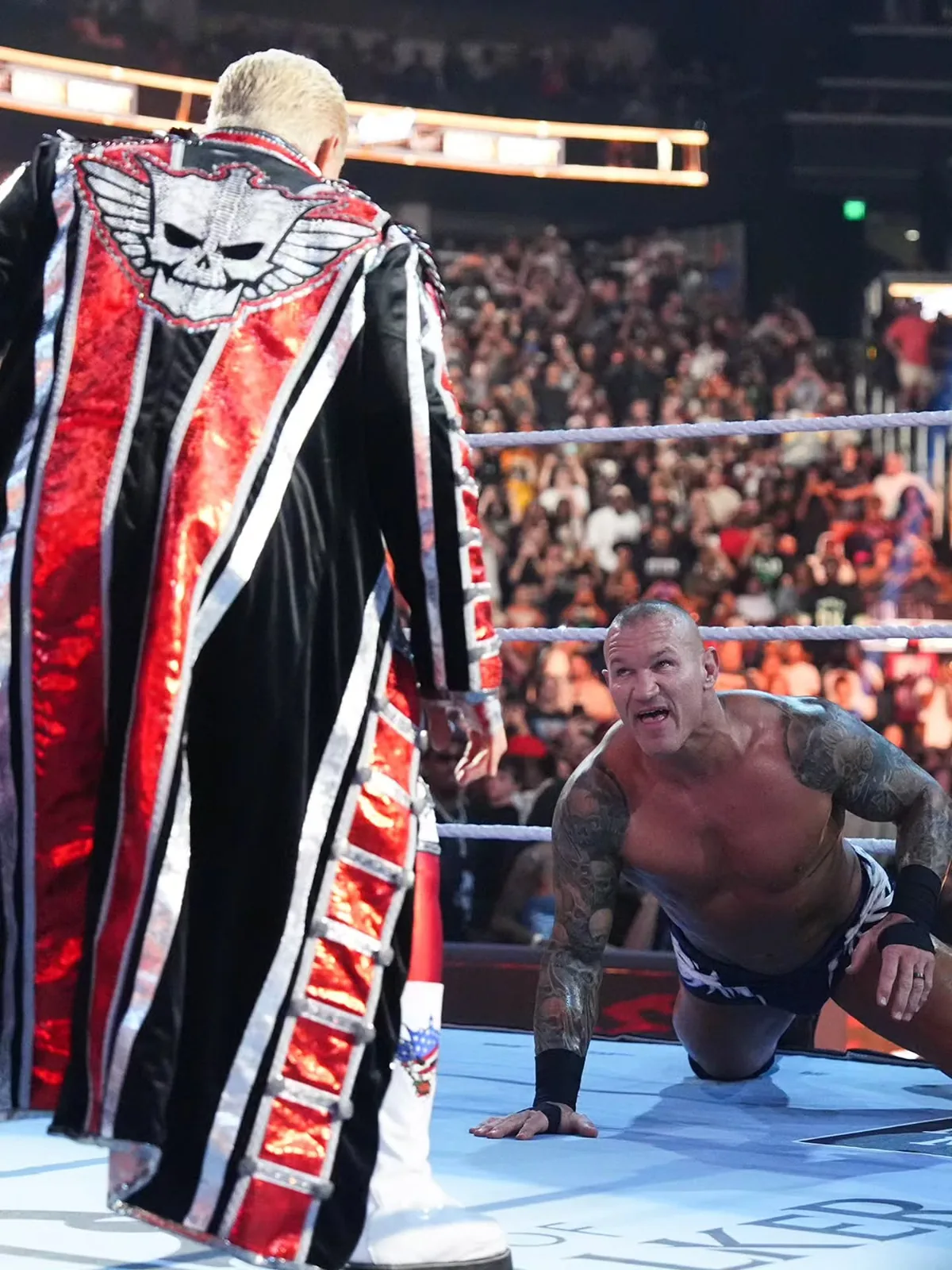 Back view of Cody Rhodes in the ring wearing the red, black, and silver entrance coat with scale shoulders
